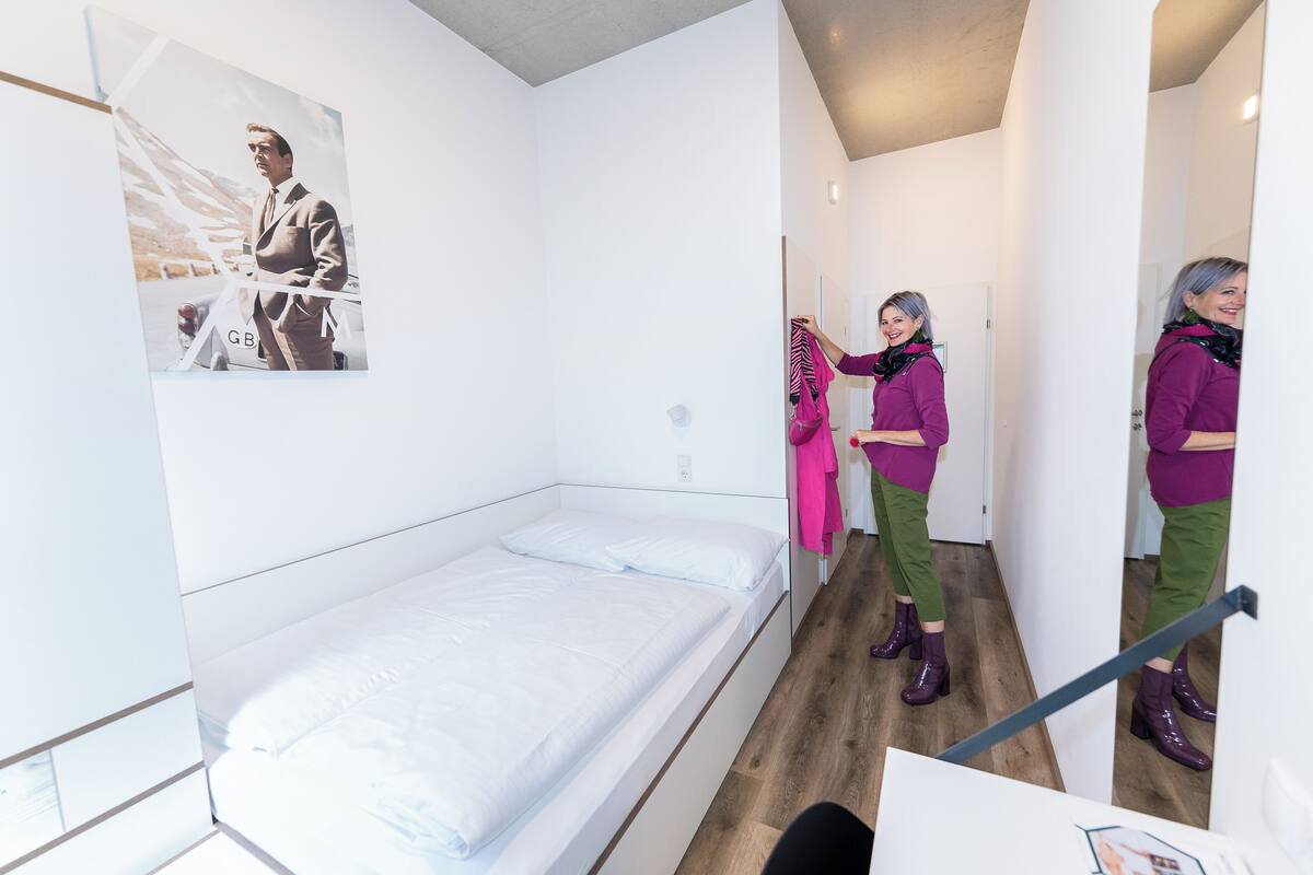 Une petite chambre moderne, avec un lit simple, un miroir et un tableau encadré au mur.