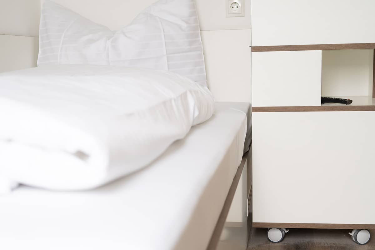 Un lit avec une literie blanche, une table de chevet avec une lampe et une prise électrique au mur.