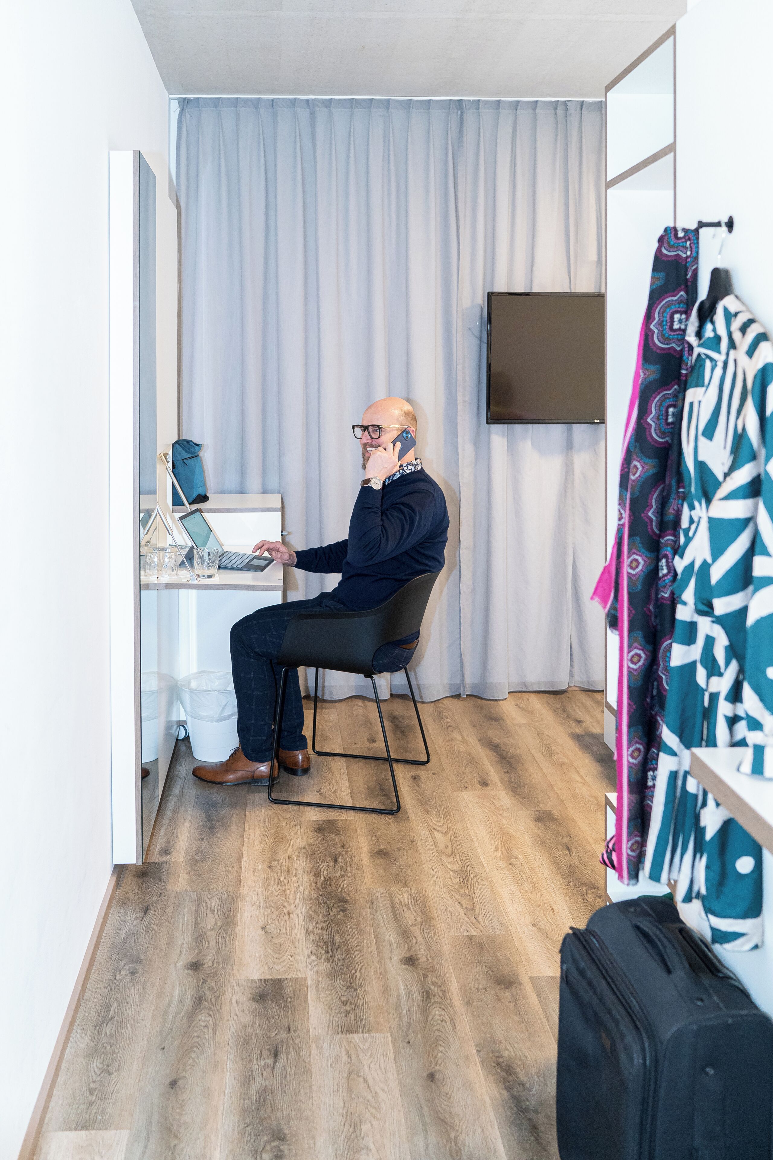 Un hombre sentado en un escritorio con una laptop, una silla y una pequeña mesa con un vaso y una taza. Hay una maleta en el piso y un perchero con un abrigo estampado.