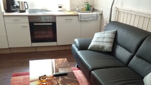 Sala de estar com sofá de couro preto, mesa de centro de madeira e cozinha ao fundo.