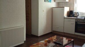 Cozinha compacta com geladeira, fogão e cafeteira. Há uma mesa pequena com tampo de vidro e um tapete vermelho. Uma porta dá acesso a outro quarto.