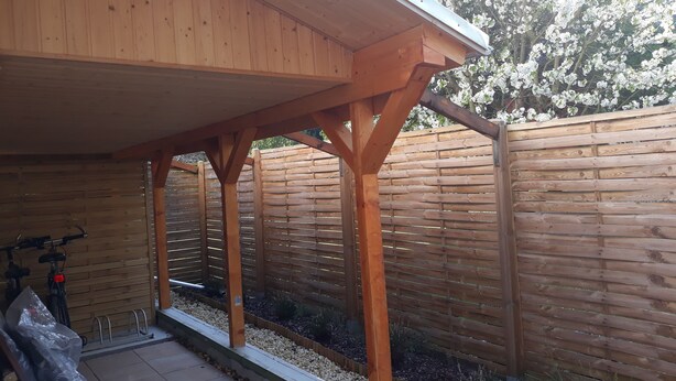 Uma estrutura de pérgola de madeira com cobertura, rodeada por um cercado de madeira e uma área de jardim com cascalho e plantas.