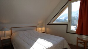 A bedroom with a sloped ceiling, a bed with white bedding, two bedside lamps, and a window with a view of mountains.