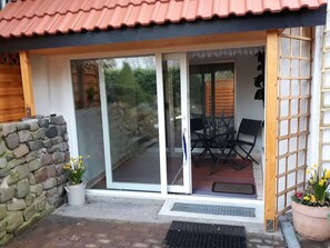 A building entrance with a stone wall, a wooden door, and a sliding glass door leading to an outdoor seating area with a table and chairs. - Vacation apartment WE 2 (Werder (Havel))