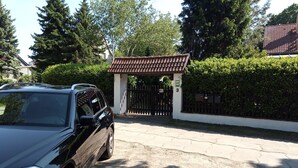 Um carro preto estacionado numa entrada de garagem com portão, cobertura em telhas e um hedge ao fundo.