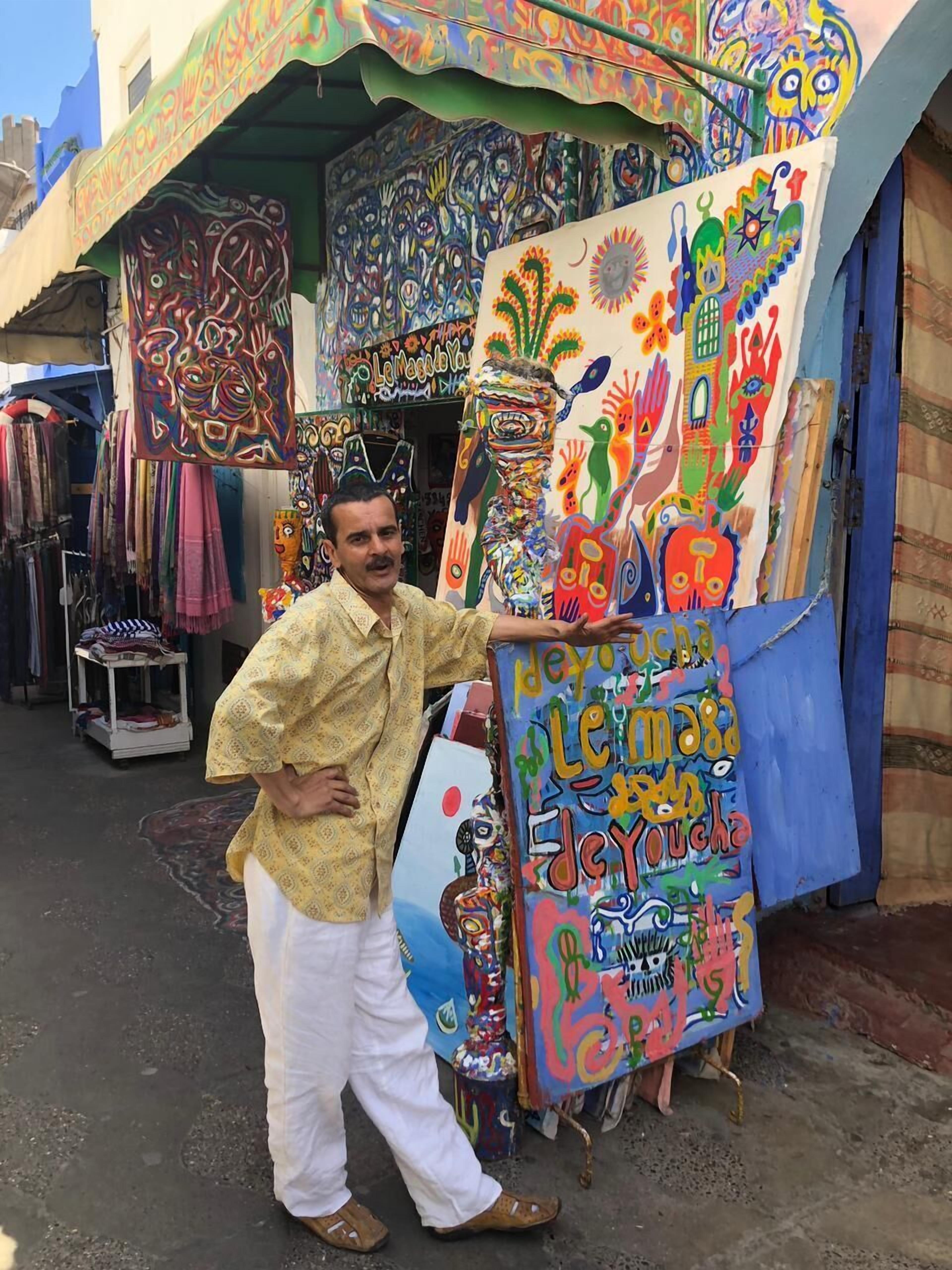 Un hombre está de pie junto a un colorido muestrario de textiles y obras de arte pintadas en un mercado al aire libre.