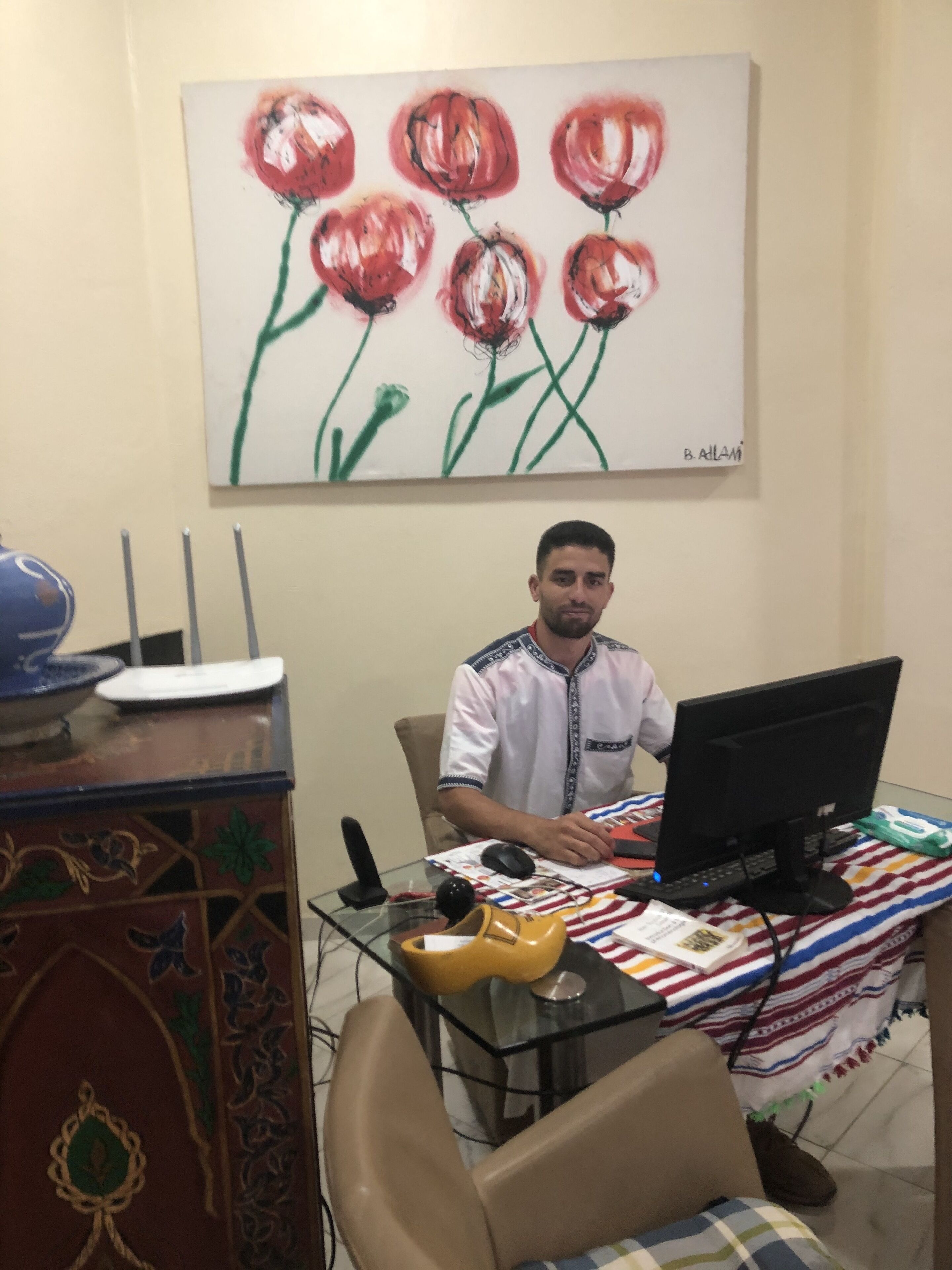 Un homme est assis à un bureau, devant un ordinateur, un téléphone et une souris. Un tableau représentant des fleurs rouges est accroché au mur derrière lui.