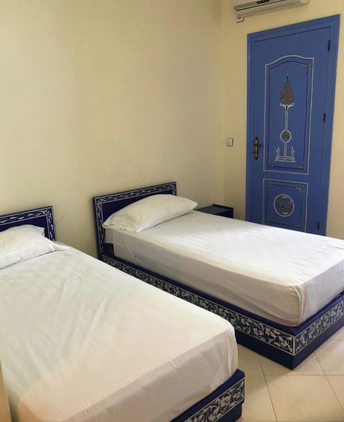 Two single beds with blue headboards and white bedding in a room with a blue door and a small air conditioning unit on the wall.