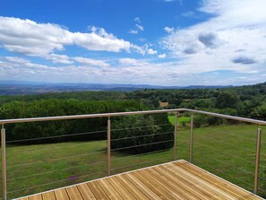 Une terrasse en bois avec une rambarde en métal, d’où l’on peut admirer un paysage verdoyant et des collines lointaines.