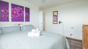 A neatly made bed with a light-colored bedspread and pillows, a wooden dresser, and two purple orchid paintings on the wall. - 4-bed Detached, Pet Friendly House in Nelson (Nelson)