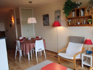 Een eetgedeelte met een tafel voor vier personen, een tafelkleed met een rood-wit patroon, witte stoelen en een rode vloerlamp.
