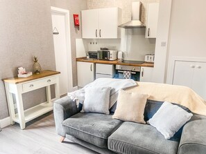 Ein kompakter Wohnbereich mit Küchenzeile, einem grauen Sofa mit gemusterten Kissen, einem kleinen Holztisch und einem weißen Schrank.