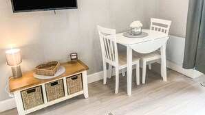 A small, neatly arranged room with a television mounted on the wall, a white table with chairs, a lamp, and wicker baskets on a wooden console. - The Rosebery Retreat By The Castle 1-bed Apartment (Inverness)