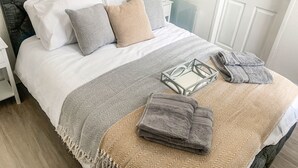 A neatly made bed with a grey headboard, white and patterned pillows, a grey throw blanket, and a glass tray with folded towels. - The Rosebery Retreat By The Castle 1-bed Apartment (Inverness)
