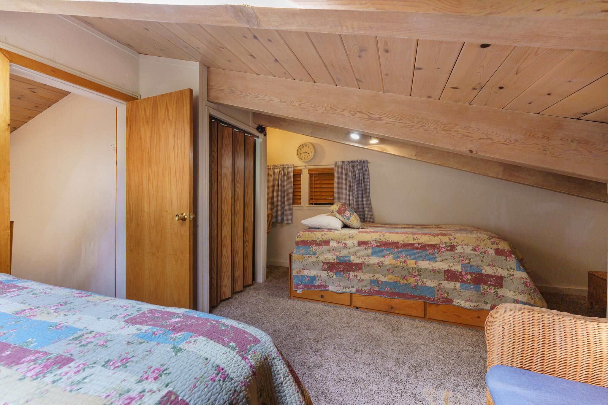 Un dormitorio acogedor en el ático con dos camas, vigas de madera, un reloj y una ventana con cortinas.
