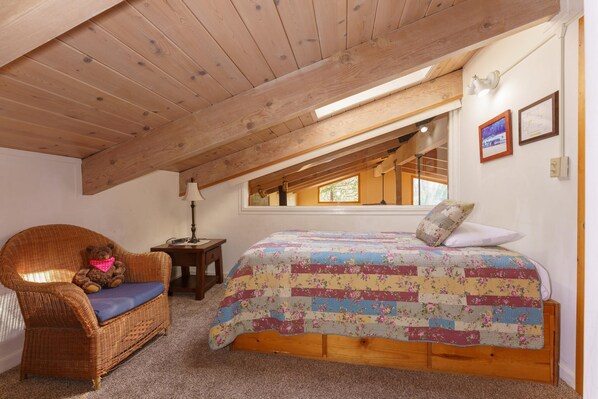 A cozy attic bedroom with a wooden ceiling, a wicker chair with a teddy bear, a quilted bed, and a window with a view.