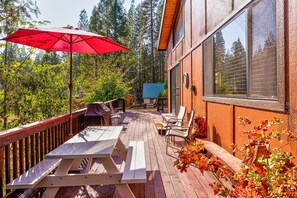 A wooden deck with picnic tables and a red umbrella, surrounded by trees and a building with large windows.