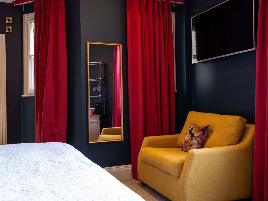 A bedroom with a bed, a yellow armchair, a large mirror, and red curtains.