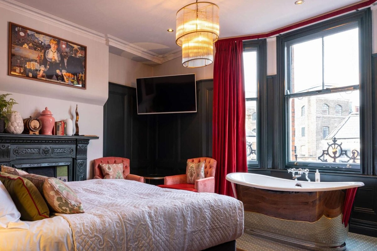 A hotel room with a large bed, a sitting area with chairs, a television, a large window with a view of buildings, and a bathtub.
