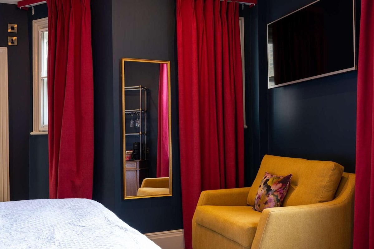 A room with a bed, a yellow armchair, a mirror, and red curtains.