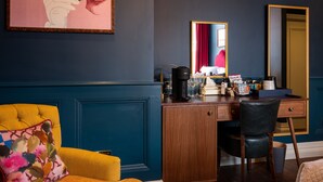 Een hotelkamer met een donkerblauwe wand, een grote spiegel, een houten bureau met een koffiezetapparaat, een stoel en een levendige gele fauteuil.