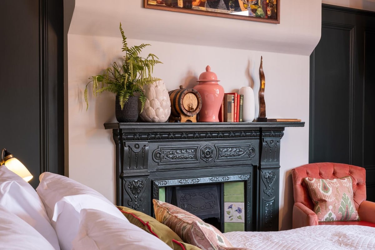 A bedroom with a bed, a chair, a fireplace with decorative items, and a wall-mounted painting.