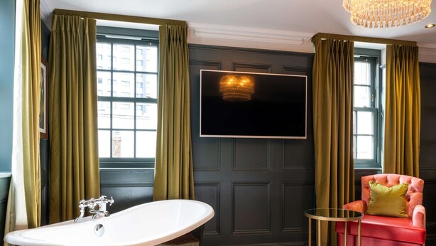 Een hotelkamer met een groot bad, een televisie bevestigd aan een donkere muur, een rode fauteuil en een kleine ronde tafel.