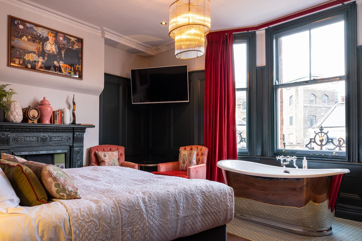 A bedroom with a large bed, a red chair, a vintage bathtub, and a large window with a view of buildings.
