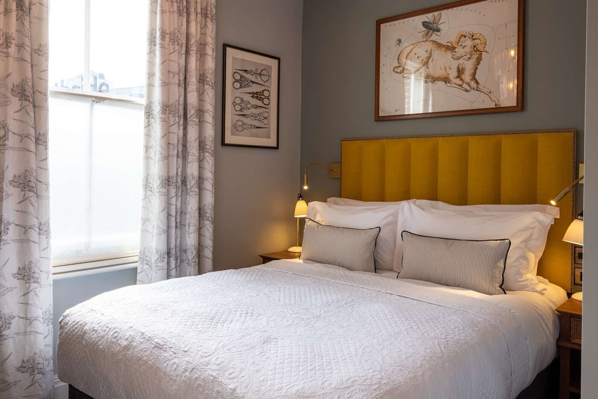 A neatly made bed with a quilted white cover, a yellow upholstered headboard, and two bedside lamps. A framed botanical illustration hangs above the bed, and a window with floral curtains is visible.