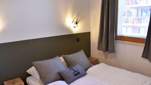 Ein ordentlich bezogenes Bett mit weißer Bettwäsche und zwei Kissen, ein kleiner Nachttisch daneben sowie eine Wandleuchte.