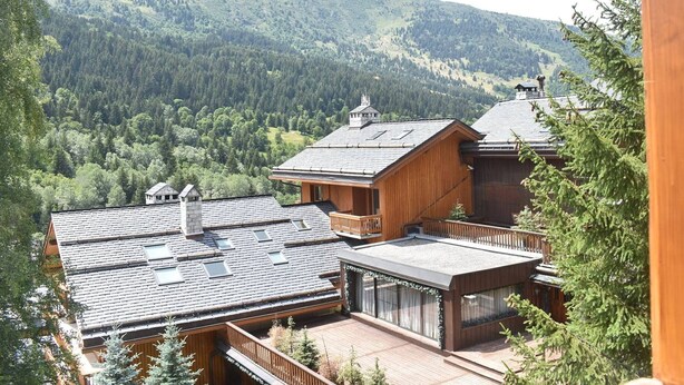 Eine hölzerne Blockhütte mit mehreren Stockwerken und großen Fenstern, umgeben von bewaldeten Bergen.