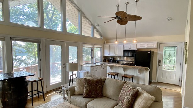 Un vaste espace de vie agrémenté d’un ventilateur de plafond, d’une grande fenêtre, d’une cuisine aux meubles blancs et d’un coin salon confortable avec un canapé et une table basse.