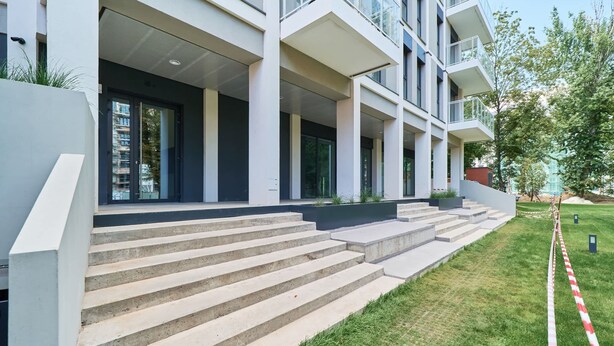Ein modernes Gebäude mit einer Treppe, die zu einem überdachten Eingang führt, einer Rasenfläche und einer roten und weißen Schutzbarriere.