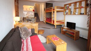 Un espace de vie compact comprenant un canapé, une petite table en bois, un téléviseur, un lit superposé et, au fond, un coin cuisine.