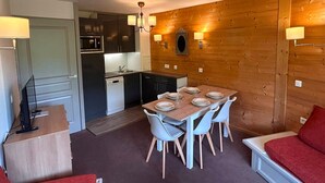 Uma cozinha compacta com eletrodomésticos modernos, sala de jantar com mesa e cadeiras de madeira, e um espaço de estar aconchegante com um sofá vermelho.