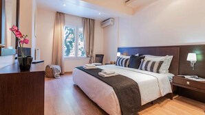 A hotel room with a bed, bedside tables, a desk, a chair, a window with curtains, an air conditioning unit, and a potted plant.