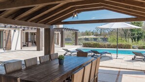 Un patio al aire libre con mesa y sillas de madera, piscina y sombrillas.
