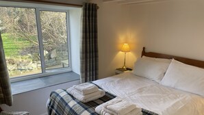 A neatly made bed with a plaid blanket and two folded towels, a bedside lamp, a chair, and a window with curtains. - Penllain (Newport)