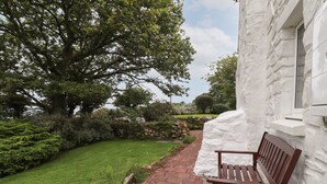A stone pathway leading to a bench in a garden with a large tree and white building. - Penllain (Newport)