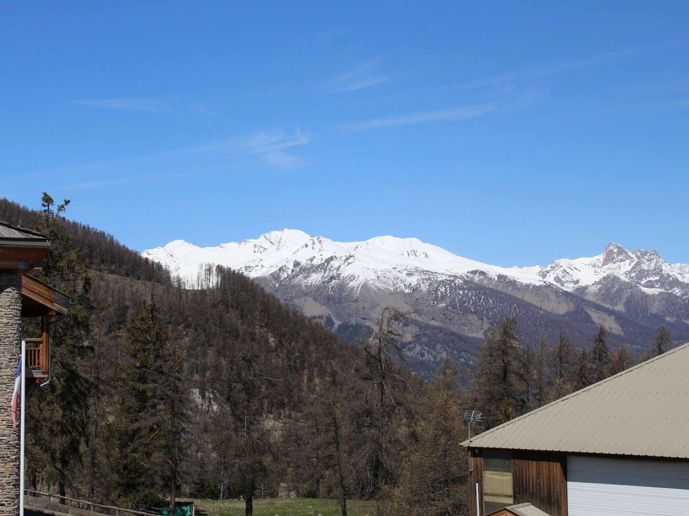 Un chalet de montagne aux murs en pierre et au toit métallique, entouré d’une forêt et de montagnes aux sommets enneigés.