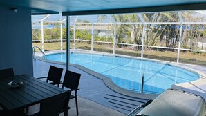 A poolside area with a table and chairs, a covered seating area, and a view of the pool and surrounding greenery.