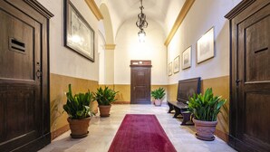Un pasillo con alfombra roja, puertas de madera, plantas en macetas e imágenes enmarcadas en las paredes.