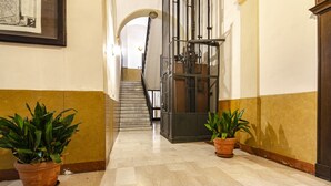 Un pasillo con suelo de mármol, dos plantas en macetas, una escalera y un ascensor de metal.