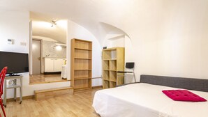 Un dormitorio con cama, escritorio y silla. Hay un espejo en la pared, y se ve una zona de cocina a través de una puerta abierta.