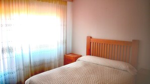 Una cama individual con cabecera de madera en una habitación con pared lisa y una ventana con cortinas translúcidas.