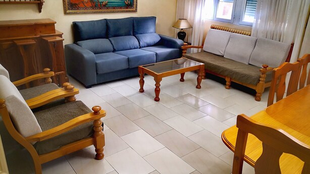 Una sala de estar con un sofá azul, sillas de madera, una mesa de centro de vidrio y una mesa de comedor de madera.