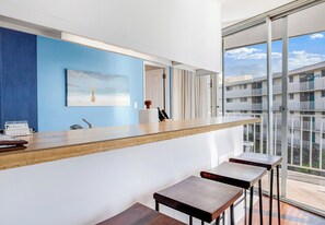 A modern bar area with a wooden counter and bar stools, a painting on the wall, and a view of a building through large windows.