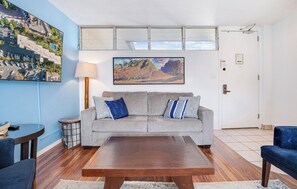 Una sala de estar con un sofá gris, una mesa de centro de madera y un cuadro de un paisaje.