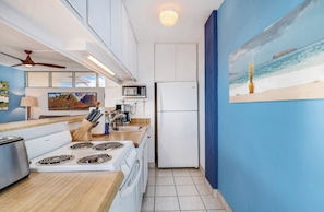 A compact kitchen with a microwave, stove, and refrigerator, a wooden countertop, and a painting of a beach scene.