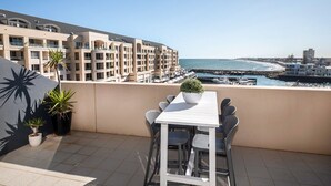 A balcony with a table and chairs, potted plants, and a view of a coastal area with buildings and a marina. - Watercolours on Marina (Glenelg)